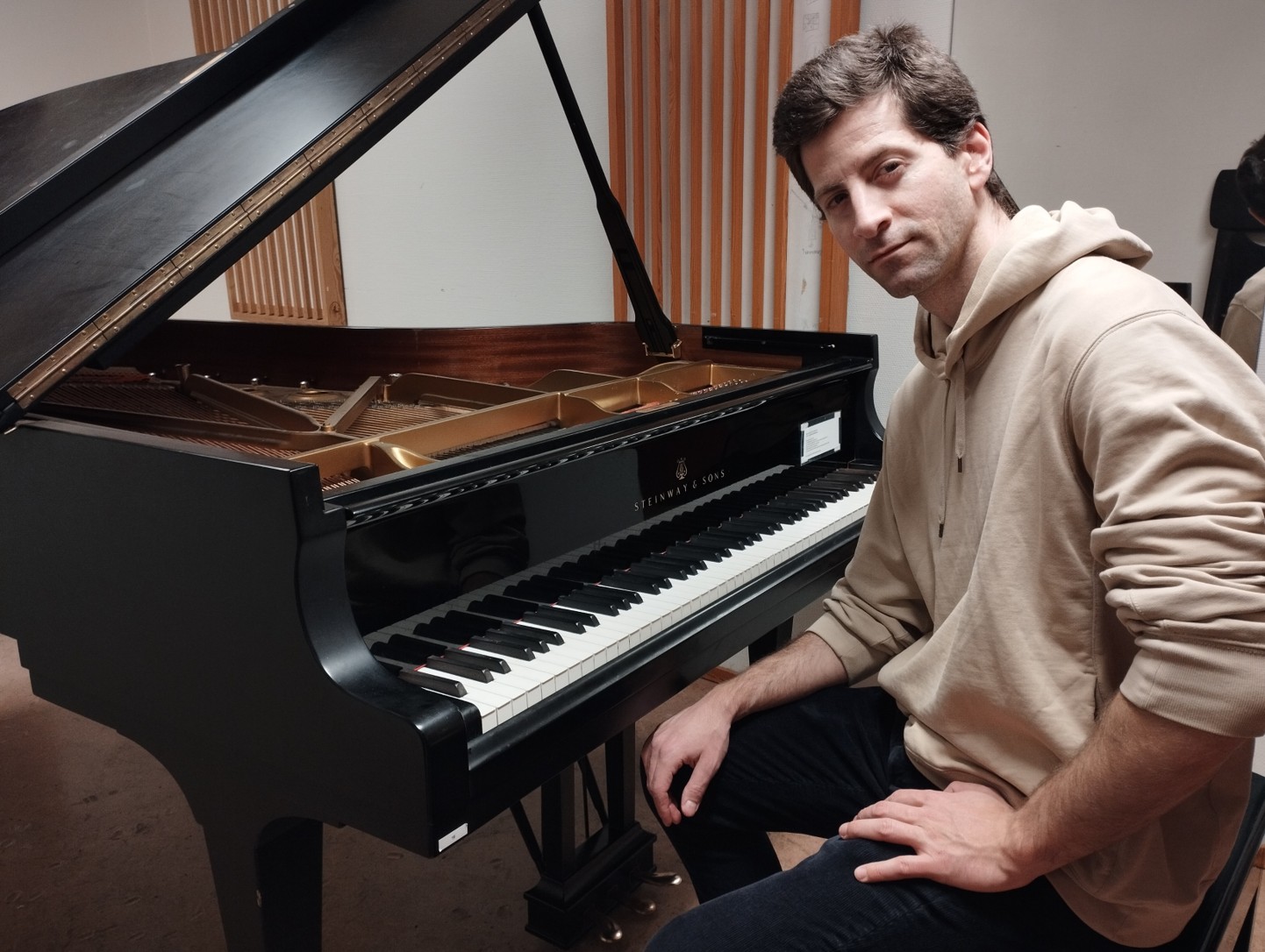 En mann i beige hettegenser sitter ved siden av et svart Steinway & Sons-flygel i et musikkrom med akustiske paneler av tre.