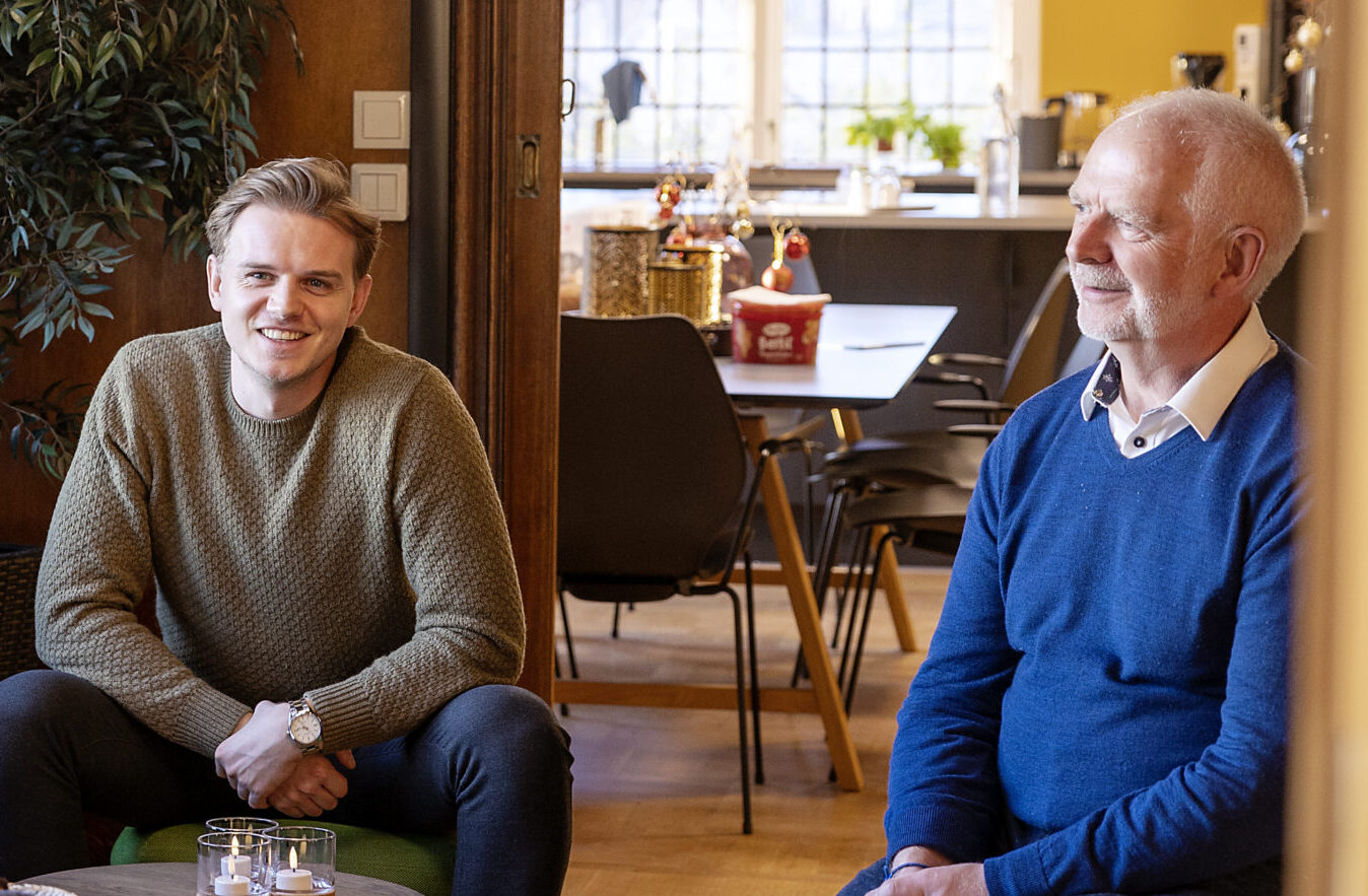 Fire personer sitter og snakker i en koselig stue med grønne sofaer, et rundt bord og store vinduer som slipper inn naturlig lys.