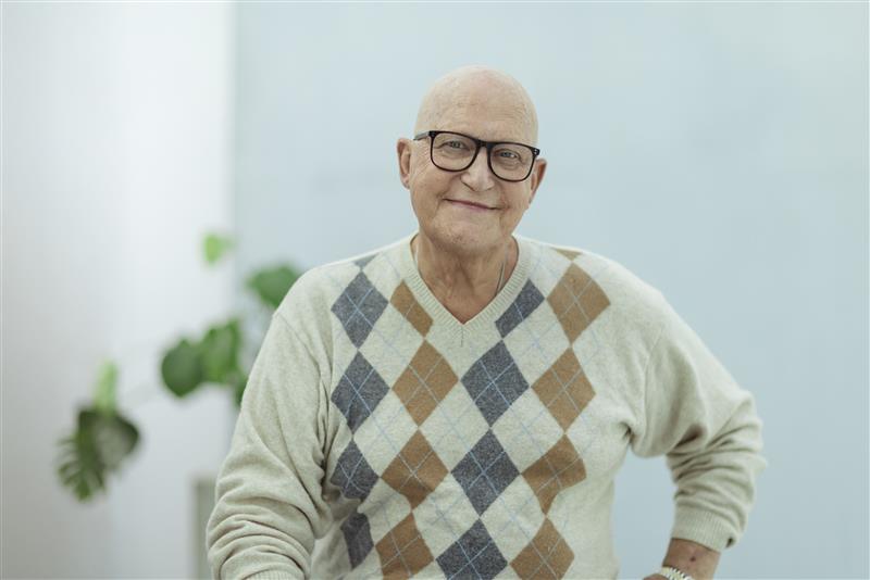 Skallet mann med briller og argylegenser smiler mens han sitter innendørs med en plante i bakgrunnen.