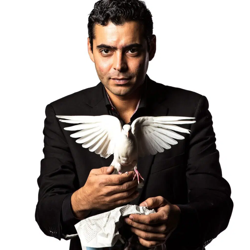 En mann i svart dress holder en hvit due med åpne vinger mot en hvit bakgrunn.