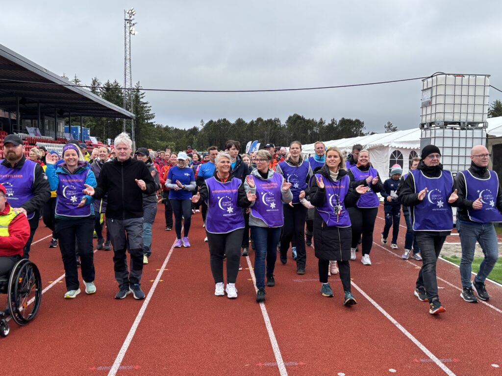 En gruppe mennesker, noen iført lilla vester, går og jogger sammen på en løpebane under et utendørsarrangement.