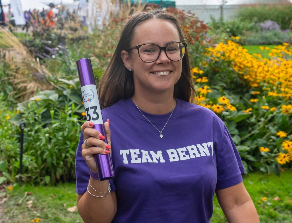 En kvinne med briller og en "TEAM BERNT"-t-skjorte holder en lilla stafettpinne med tallet 33, mens hun står i en hage med gule blomster.