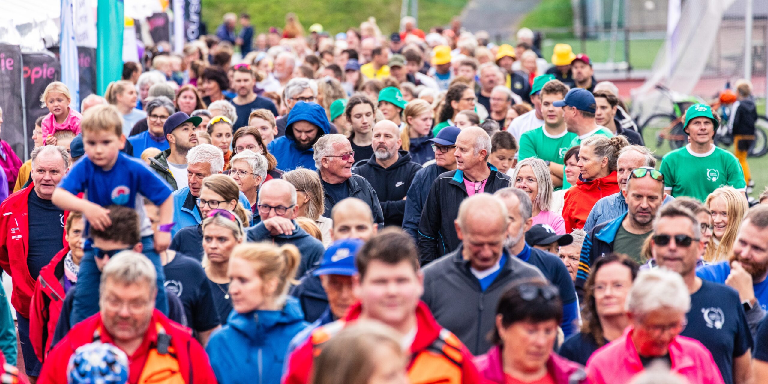 En stor folkemengde, inkludert voksne og barn, går utendørs på et organisert offentlig arrangement, mange iført fargerike klær.