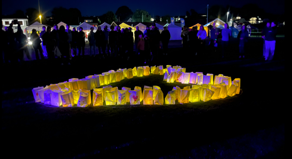 En gruppe mennesker står rundt lysende luminariaposer som er plassert i en stor trekant på gresset i mørket.