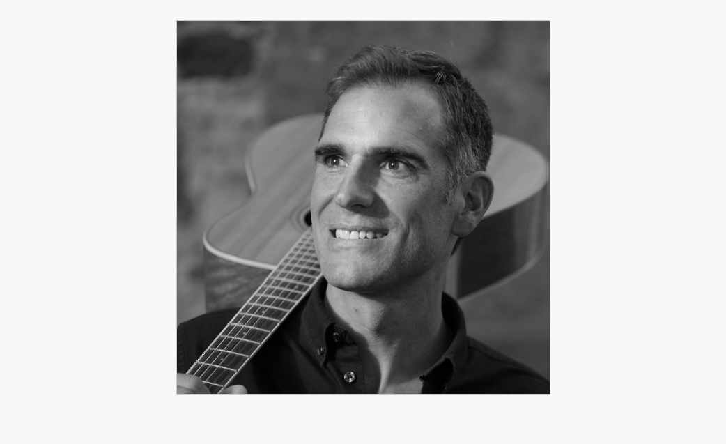 Svart-hvitt-foto av en mann som smiler og holder en akustisk gitar over skulderen, mens han ser litt til venstre.