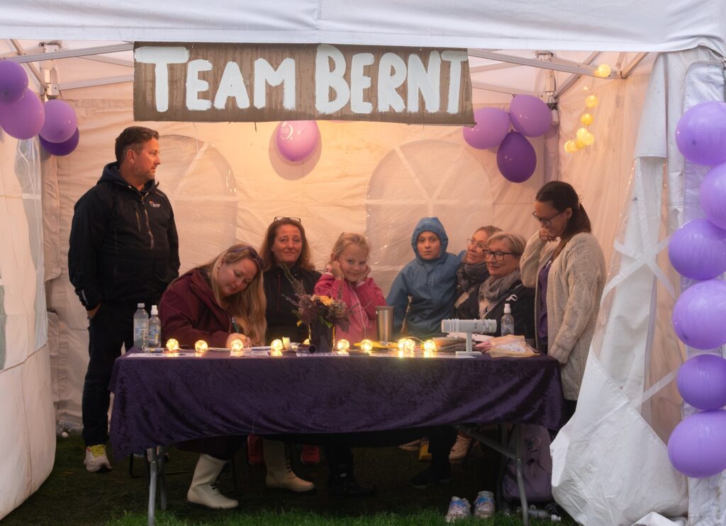 En gruppe mennesker sitter og står under et telt dekorert med lilla ballonger og et "TEAM BERNT"-skilt, samlet rundt et bord med lys og blomster.