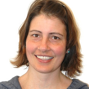 Kvinne med kort, brunt hår og blå topp smiler til kameraet mot en vanlig, hvit bakgrunn.