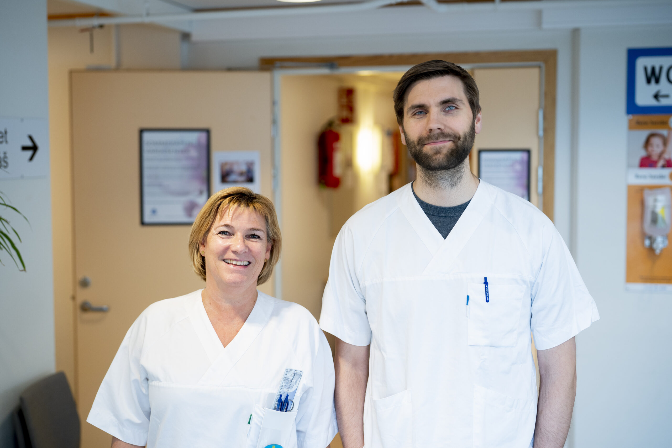 To helsepersonell i hvite uniformer står side om side i en sykehuskorridor og smiler til kameraet.