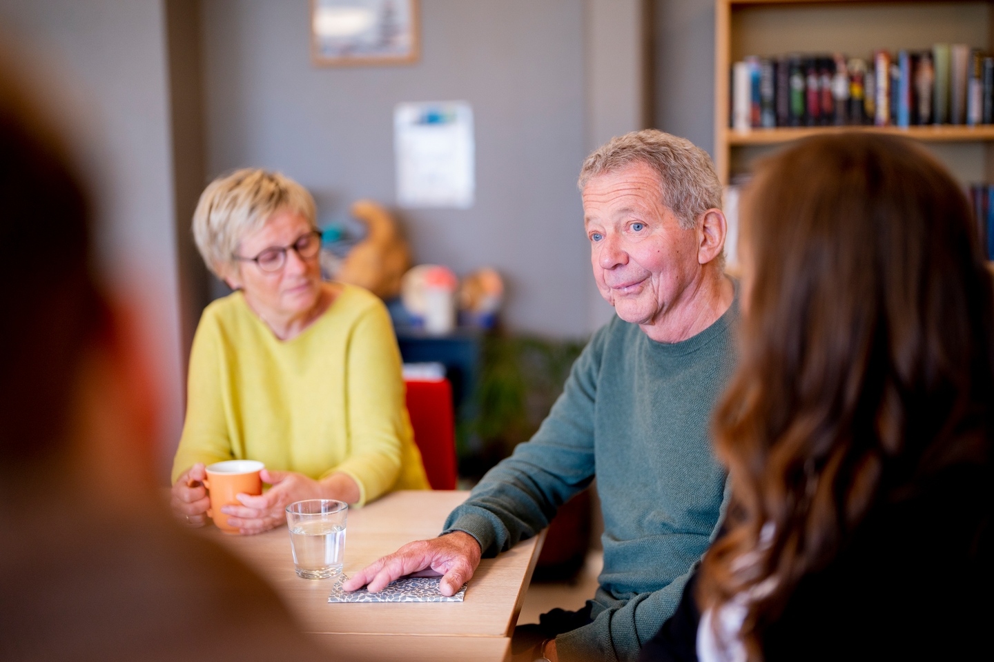 Fire personer sitter rundt et bord og samtaler. En eldre mann snakker, mens en eldre kvinne med briller lytter. Bokhyller er synlige i bakgrunnen.