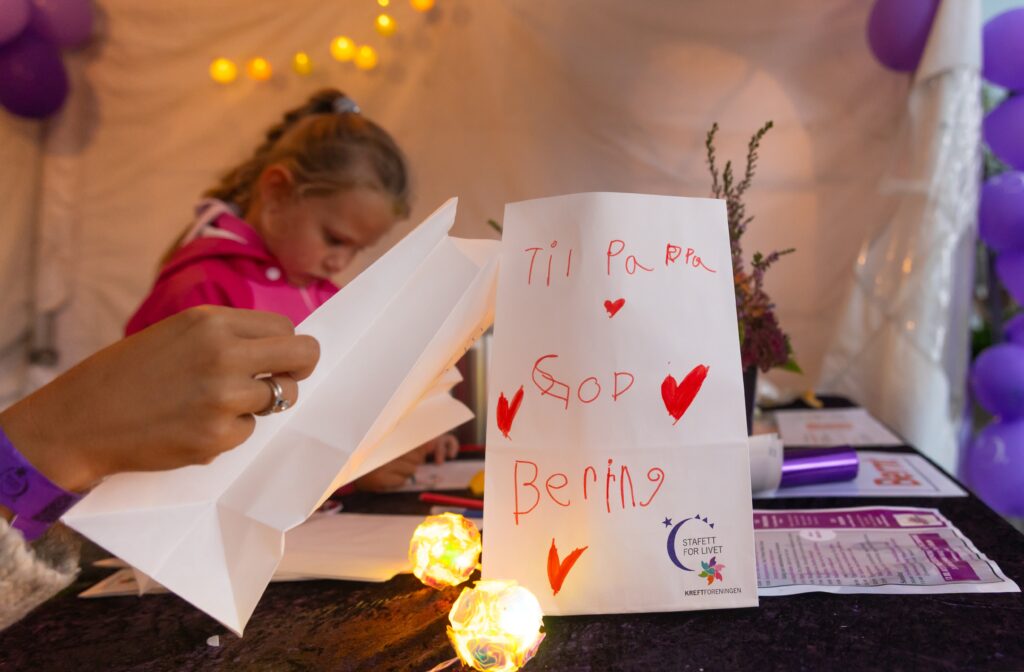 Et barn pusler ved et pyntet bord med en papirlykt med teksten "Til Pappa God Bedring" blant lilla ballonger og lyskastere.