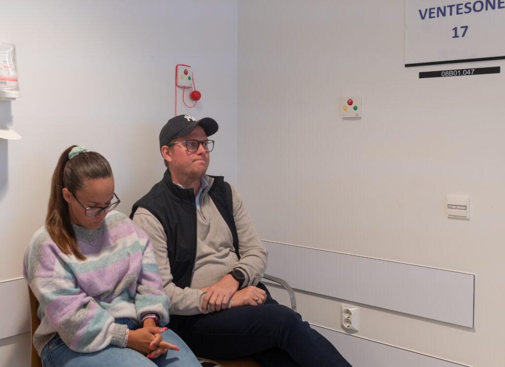 To personer sitter i et venterom; den ene ser ned på en telefon, mens den andre ser fremover med hendene foldet i fanget. På veggen henger et skilt med "VENTESONE".