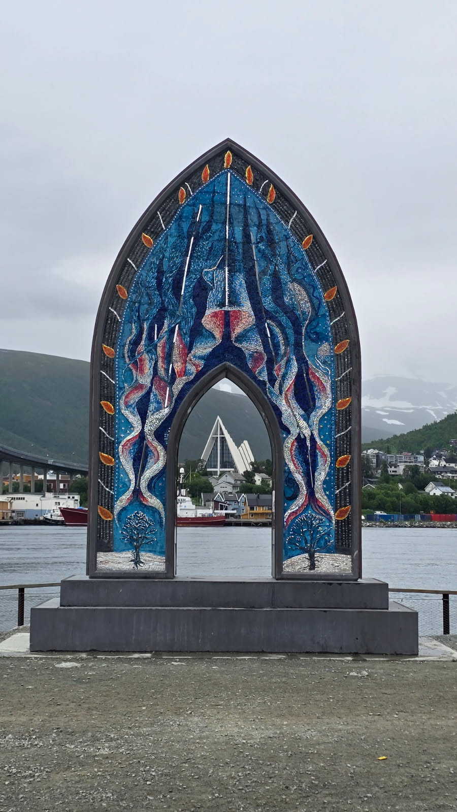 En fargerik bueskulptur i mosaikk står ved en vannkant og rammer inn utsikten mot en spiss hvit bygning og åser i bakgrunnen under en skyet himmel.