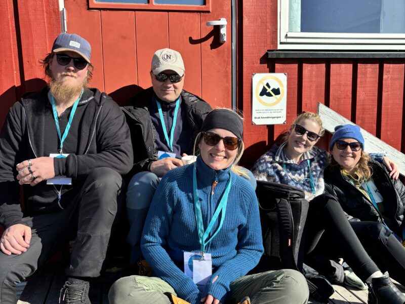 Fem personer iført utendørsklær sitter på et tredekke foran en rød bygning og smiler til kameraet. Et skilt med en svart og gul logo er synlig på veggen ved siden av dem.