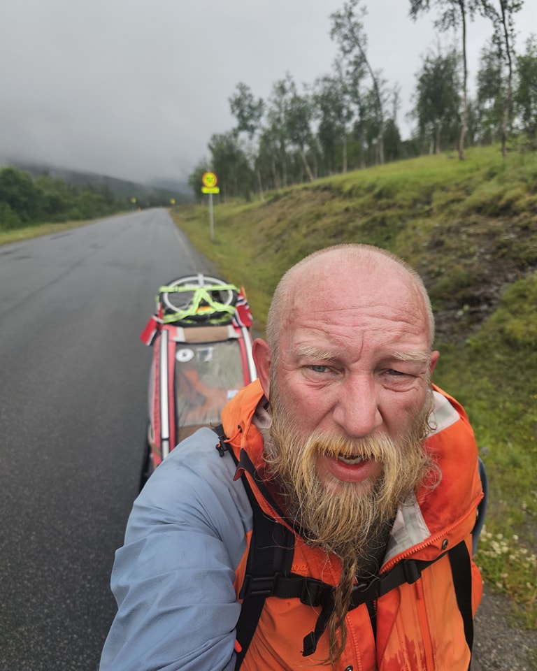 En skjeggete person i oransje jakke går langs en våt vei og trekker en kjerre, med trær og tåkete åser i bakgrunnen.