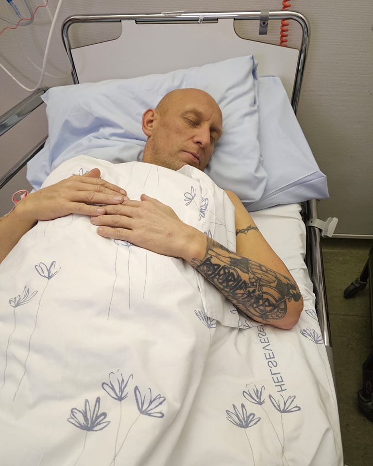 En skallet person med en tatovert arm ligger i en sykehusseng med lukkede øyne, dekket av et hvitt, blomstermønstret teppe.