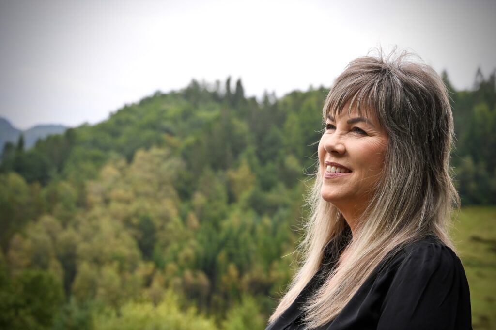 En kvinne med langt grått hår og svart topp står utendørs og smiler, med en bakgrunn av grønne trær og åser under en skyet himmel.