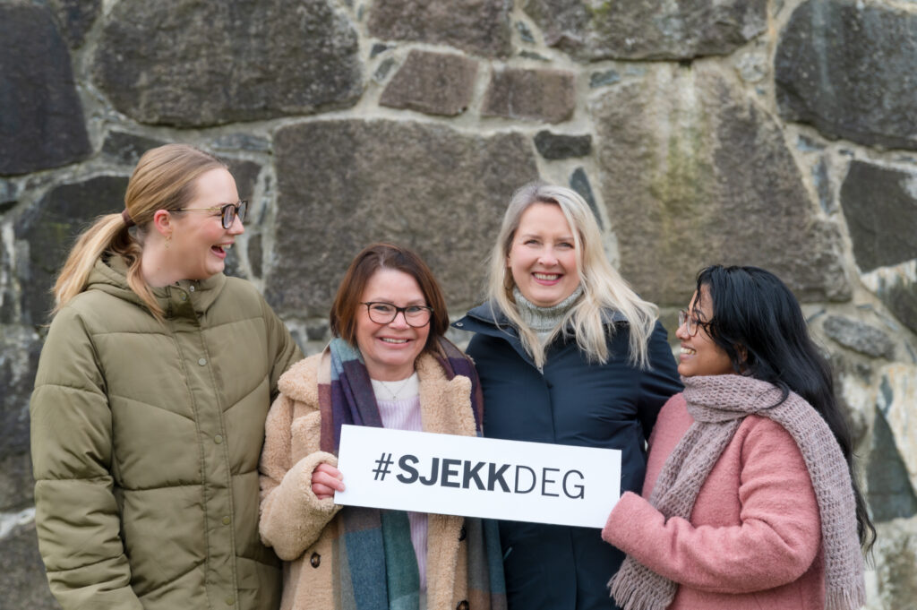 Fire vinterkledde kvinner står smilende foran en steinmur, mens en av dem holder et skilt med teksten "#SJEKKDEG".