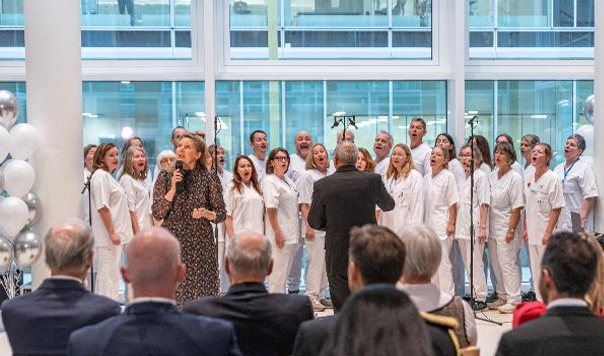 Et hvitkledd kor opptrer innendørs under ledelse av en dirigent, mens en kvinne står foran med en mikrofon. Et publikum sitter i forgrunnen.