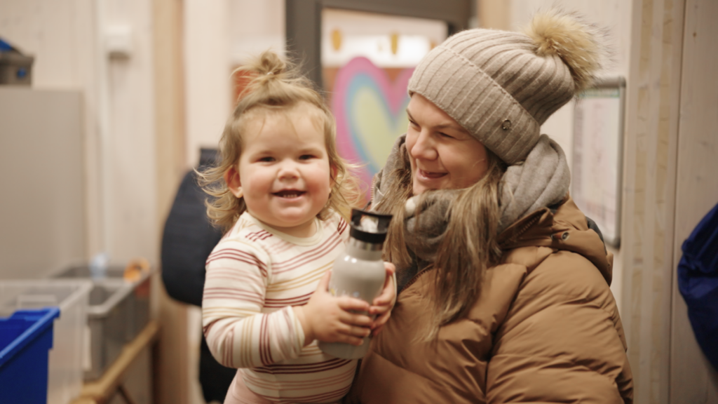 En kvinne i brun vinterfrakk og strikkelue holder et smilende barn i stripete pysjamas som holder i en vannflaske. Innendørs, med fargerike kunstverk i bakgrunnen.