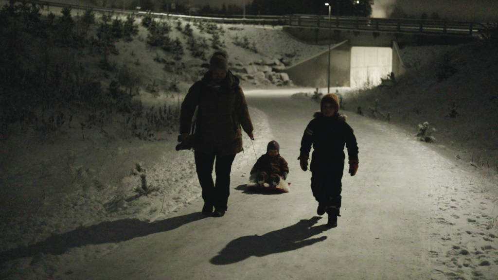 Tre personer går på en snødekt sti om natten; en voksen og et barn går, mens et annet barn sitter på en kjelke. En bro og trær er synlige i bakgrunnen.