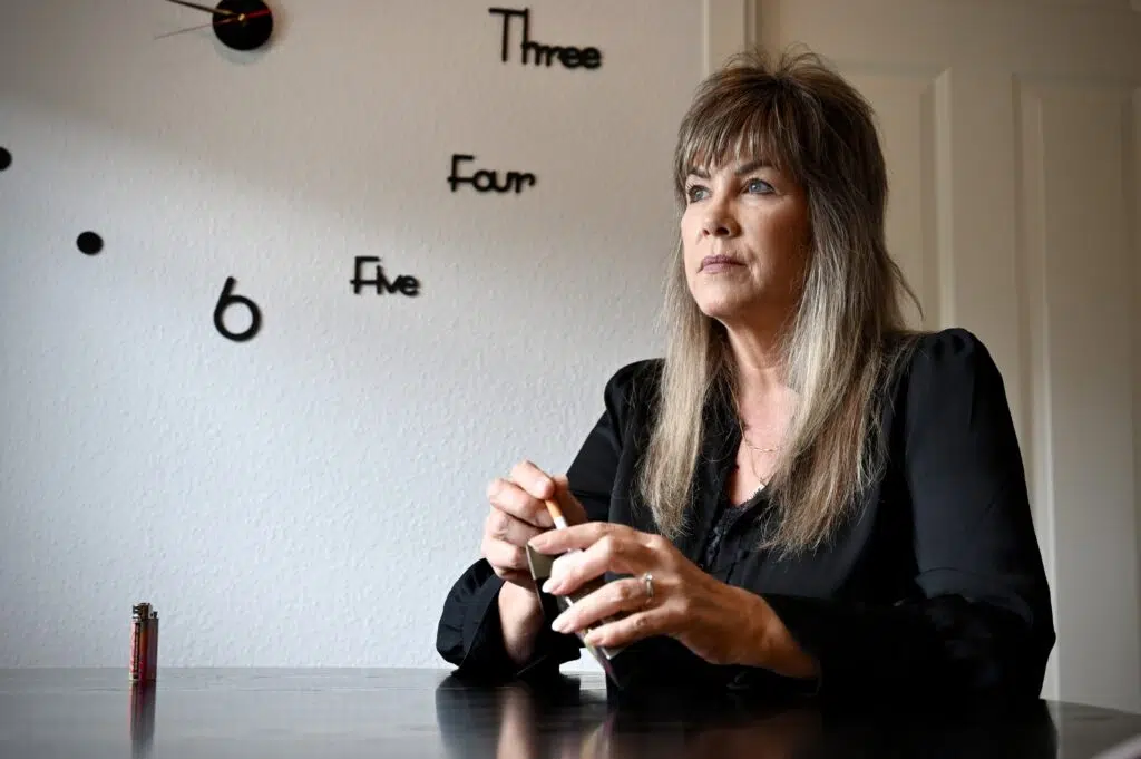 En kvinne med langt hår sitter ved et bord med batterier i hånden, og bak henne henger en veggklokke med tall skrevet som ord.