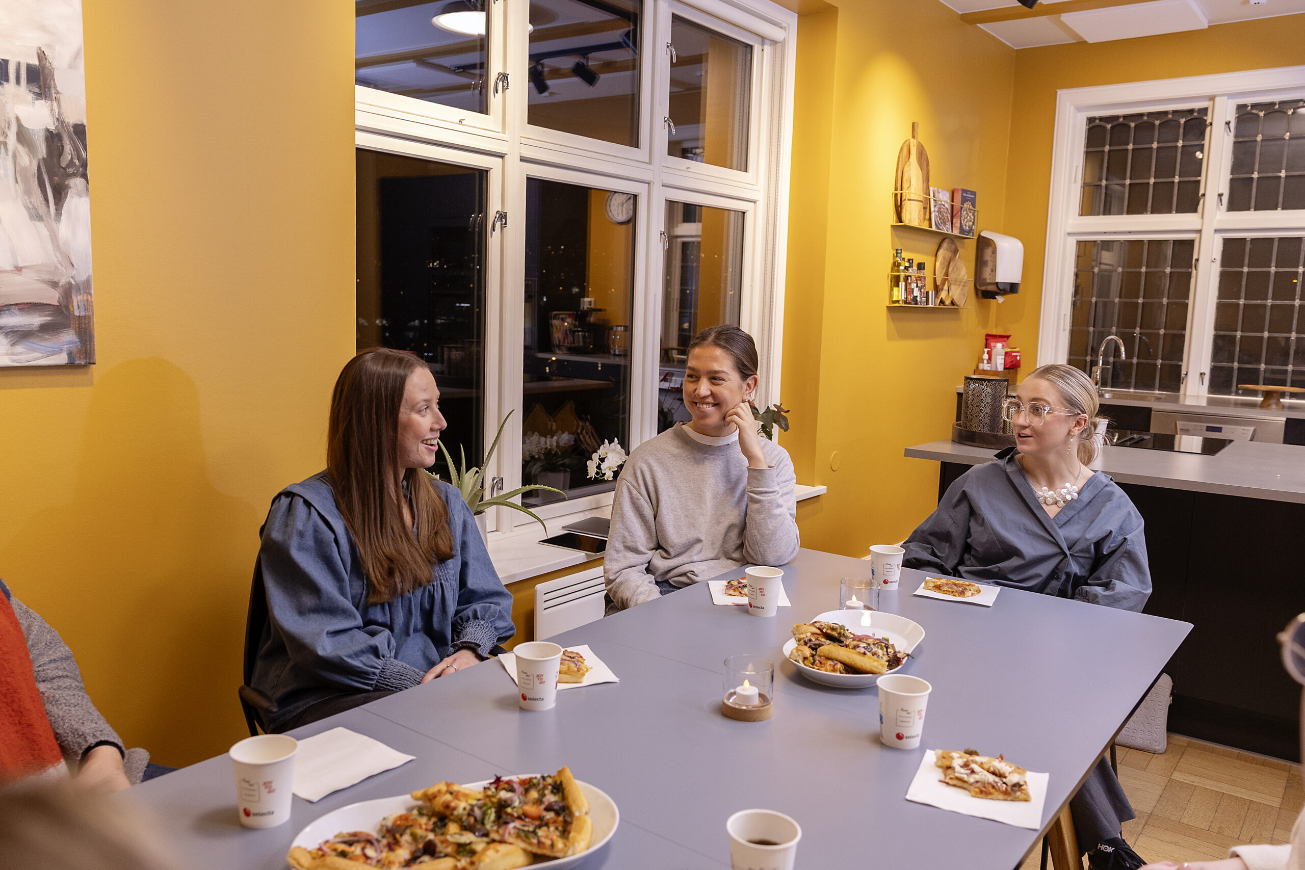 Tre personer sitter rundt et bord med pizza og drikke i et lyst rom og snakker sammen.