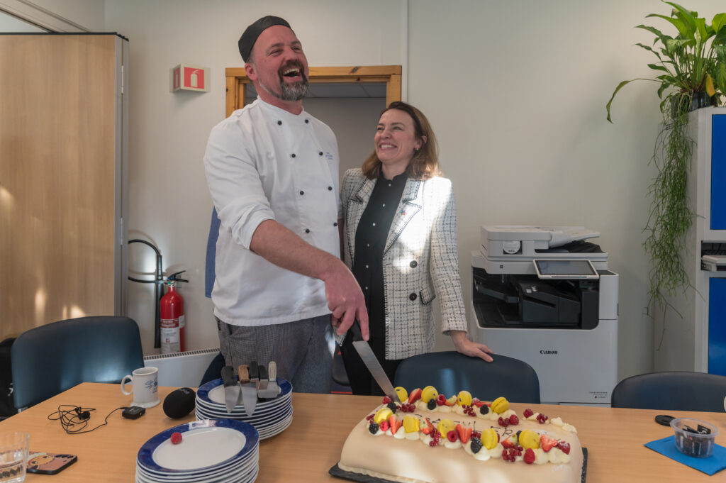 En mann i kokkeuniform og en kvinne står ved et bord mens mannen skjærer opp en kake med frukt på toppen. Begge smiler. Tallerkener, kopper og bestikk står på bordet i et kontormiljø.