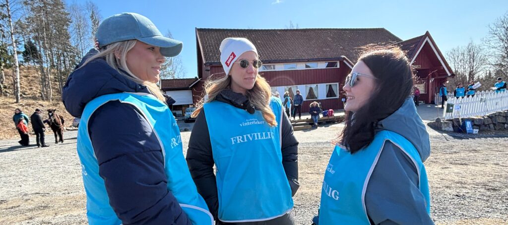 Tre kvinner iført blå dugnadsvester og vinterklær står og snakker utendørs i nærheten av en rød bygning på en solrik dag.