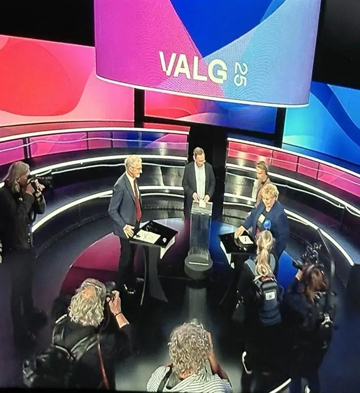 Folk deltar i en TV-sendt debatt eller diskusjon, med kameraer og fotografer til stede, under en storskjerm som viser "VALG 23".