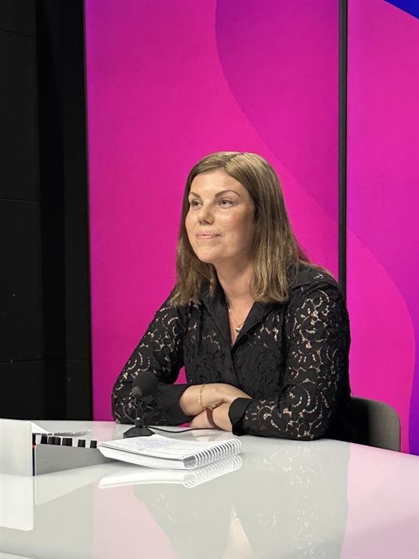 En kvinne med brunt hår sitter ved et skrivebord med mikrofon og notatbok, foran en rosa og lilla bakgrunn.