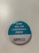 En rund blå og hvit knapp med norsk tekst som promoterer livmorhalsscreening, der det står "Spør meg om livmorhalsprøve" og "Sjekk deg Kreftforeningen.".