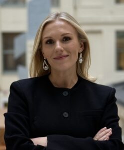 En kvinne med langt, blondt hår, svart blazer og øreringer står innendørs med armene i kors og ser inn i kameraet.