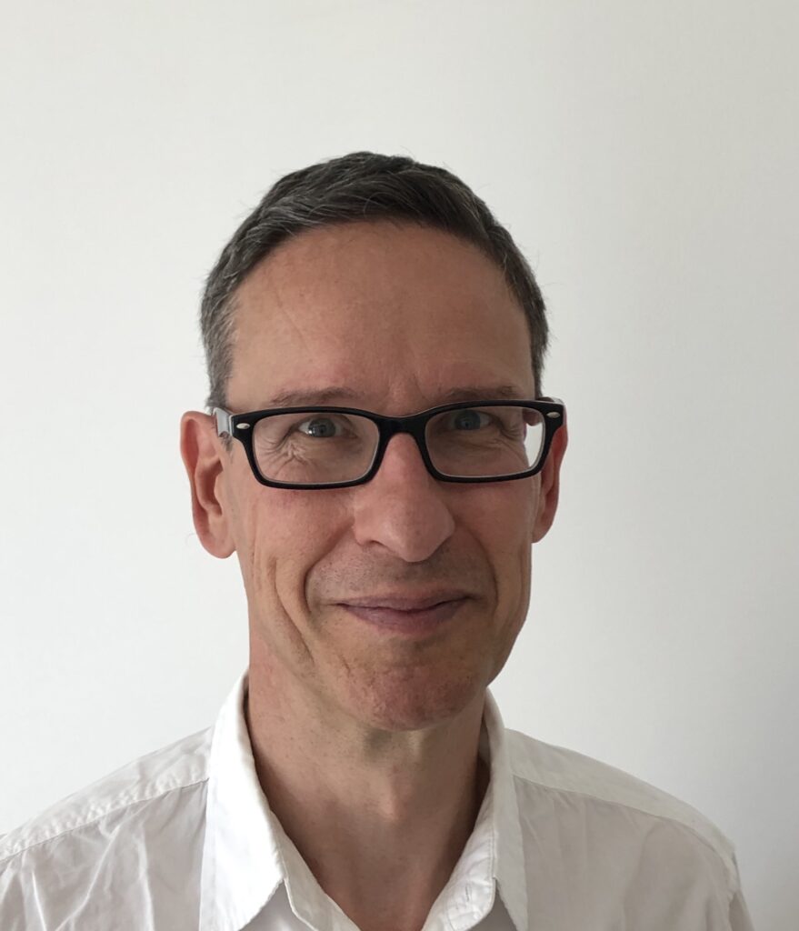 En middelaldrende mann med kort, grått hår og svarte briller, iført hvit skjorte med krage, står foran en vanlig hvit bakgrunn.