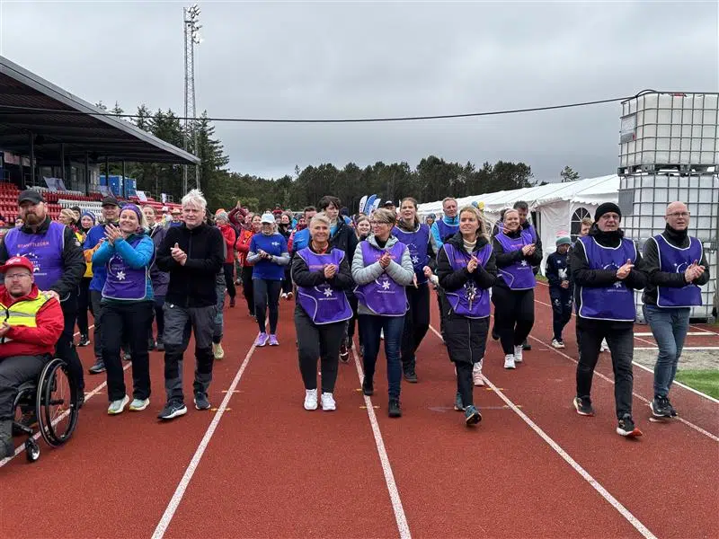 En gruppe mennesker, noen i lilla vester, går sammen på en løpebane under et utendørsarrangement; flere klapper, og en person i rullestol er til stede.