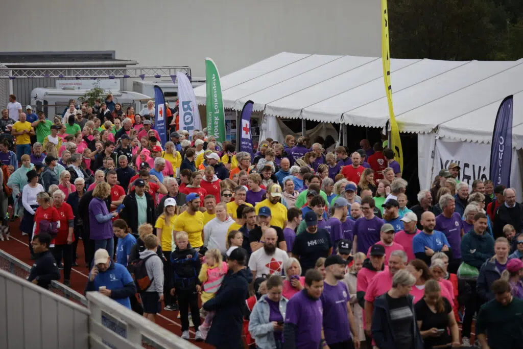 En stor folkemengde iført fargerike skjorter går på en bane ved siden av et hvitt arrangementstelt med ulike bannere og flagg.