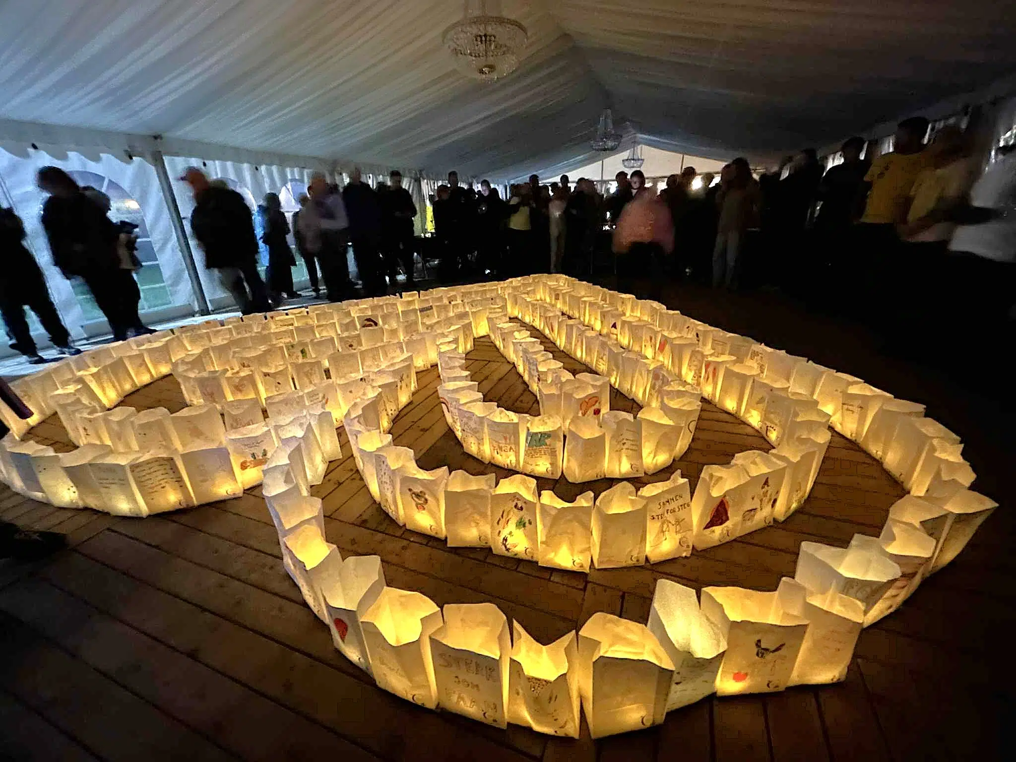 Papirlykter i et spiralmønster på gulvet lyser opp mens folk står og ser på inne i et teltområde.