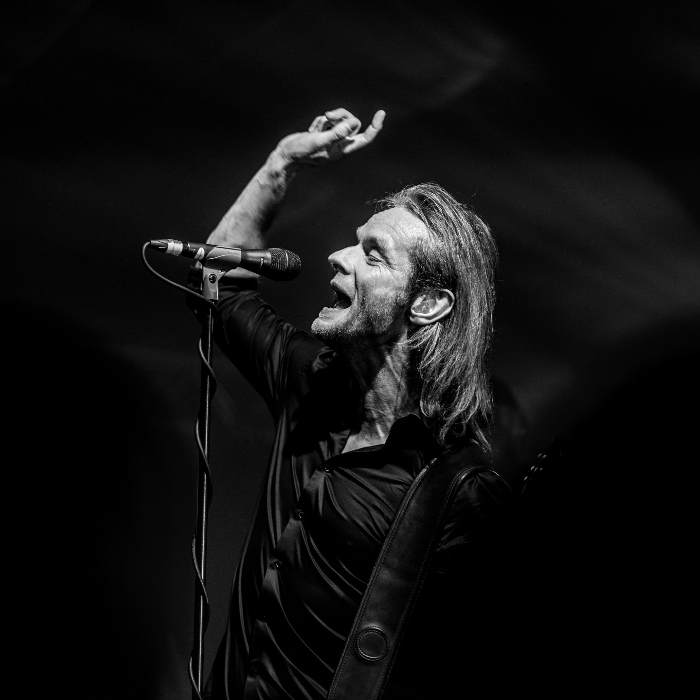 En mann med langt hår synger lidenskapelig inn i en mikrofon på scenen, mens han løfter den ene armen, i et svart-hvitt foto.