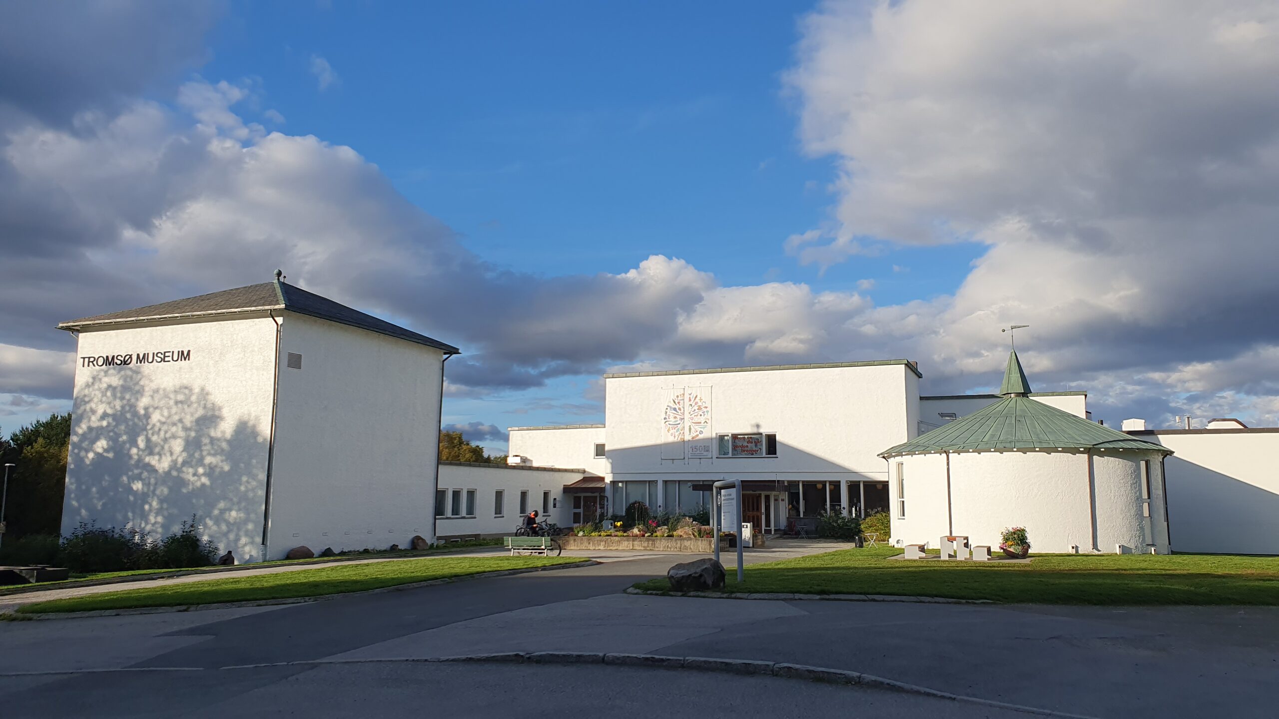 Tromsø Museums hvite bygninger under en delvis overskyet blå himmel, med en liten bygning med grønt tak til høyre og en asfaltert innkjørsel i forgrunnen.