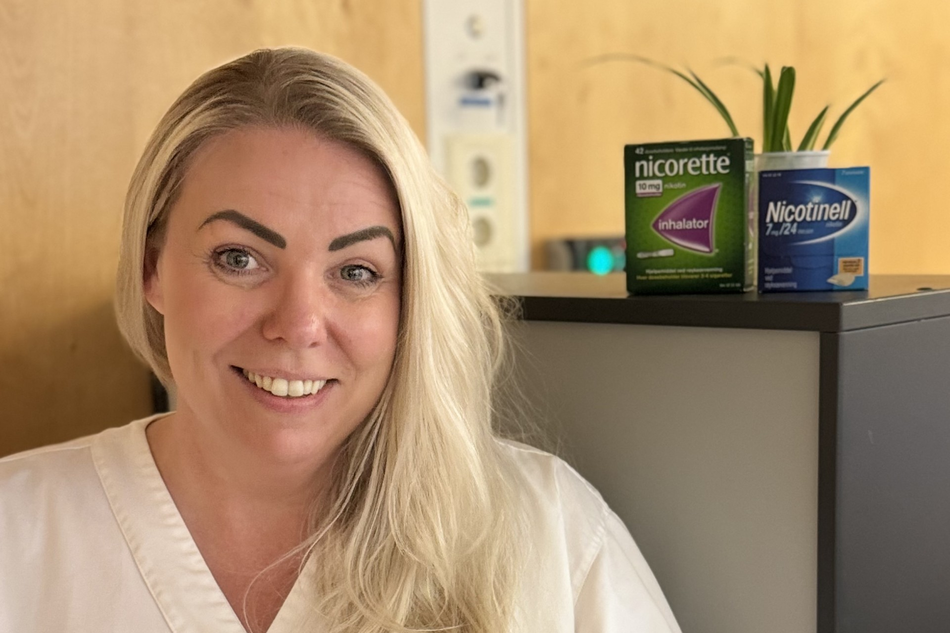 En kvinne i hvit legeuniform står ved siden av et skap med Nicorette- og Nicotinell-produkter og en liten potteplante på toppen.
