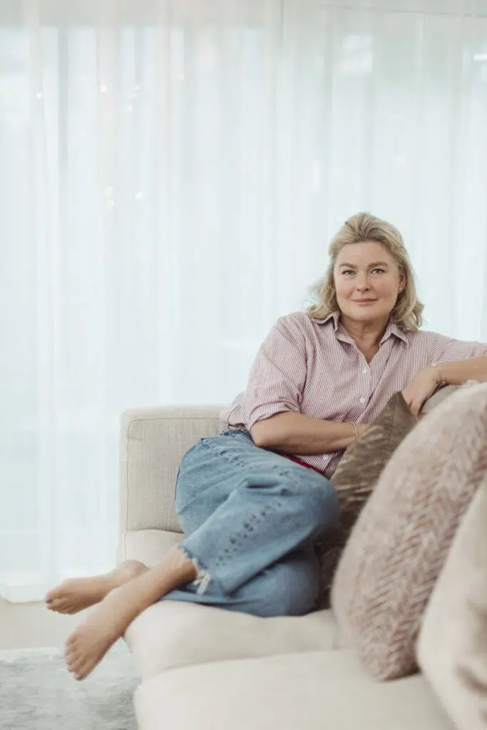 En kvinne sitter barbeint i en lys sofa, iført stripete skjorte og jeans, med armen hvilende på en pute og skira gardiner i bakgrunnen.