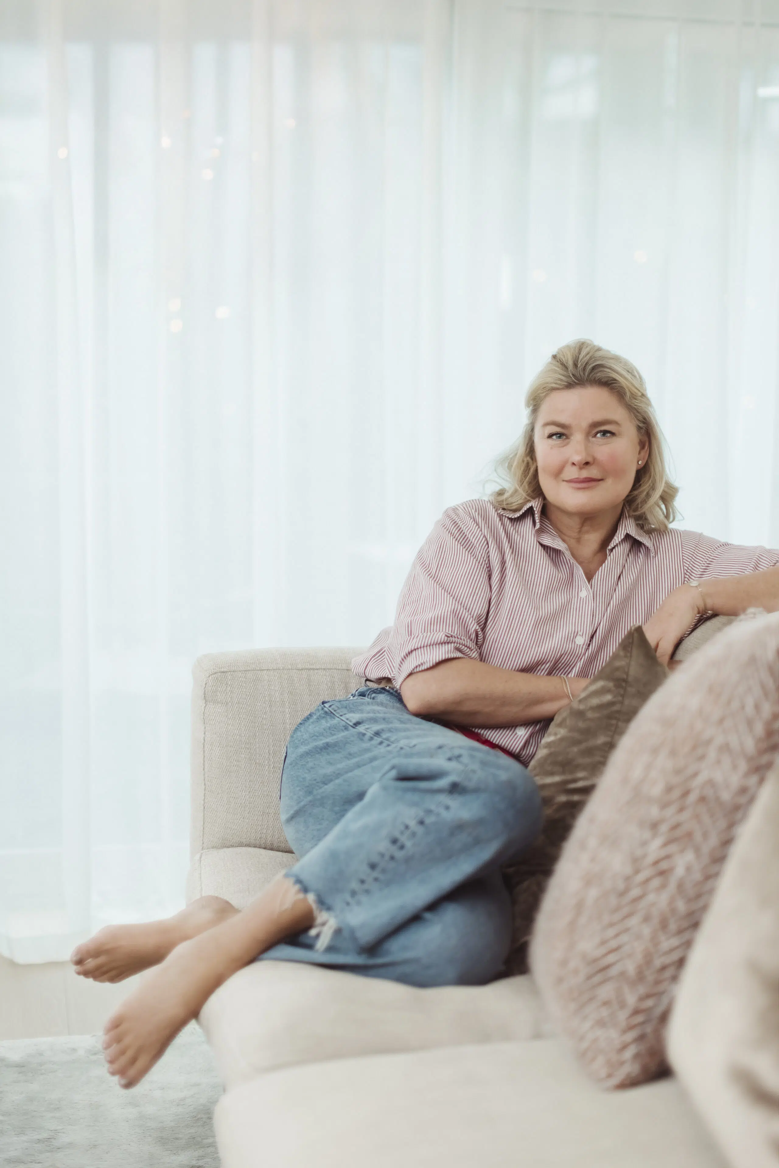 En kvinne sitter barbeint i en lys sofa, iført stripete skjorte og jeans, med armen hvilende på en pute og skira gardiner i bakgrunnen.