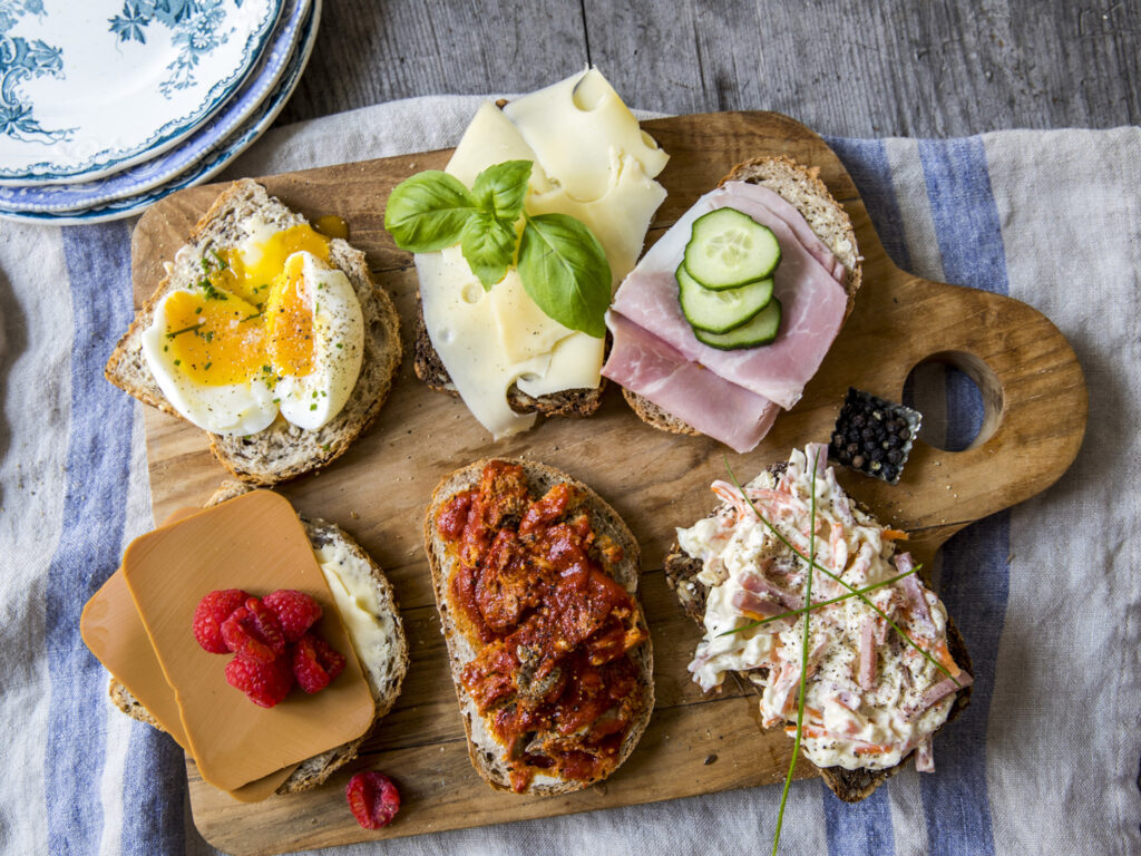 Et trebrett med seks smørbrød med ulike pålegg, blant annet egg, ost, skinke, agurk, bringebær, tomat og kremet coleslaw.