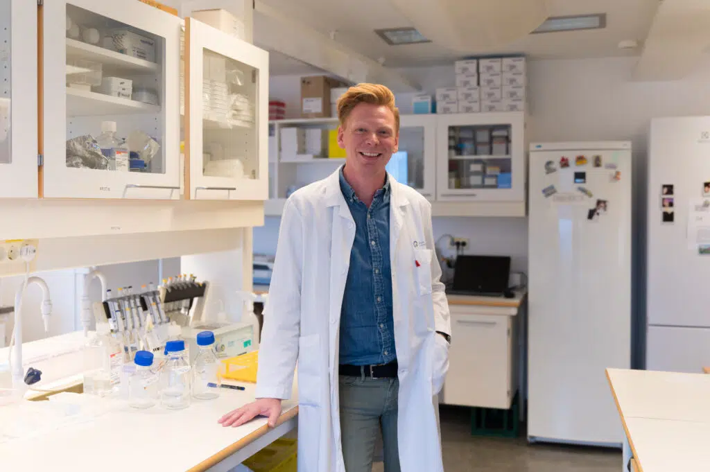 En person i hvit laboratoriefrakk står og smiler i et laboratorium, omgitt av vitenskapelig utstyr og rekvisita.