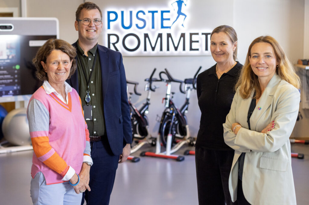 Fire personer står foran stasjonære sykler og et skilt med teksten "PUSTEROMMET" i en treningsstudio-lignende setting.