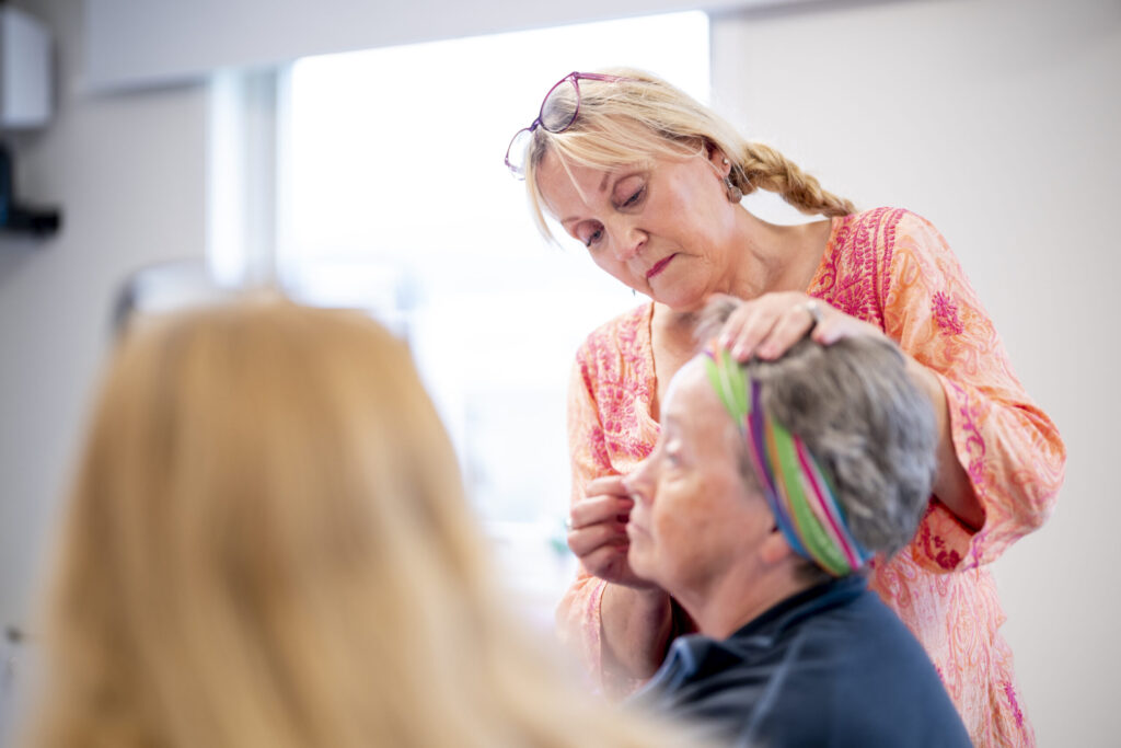 En kvinne sminker en annen kvinne som sitter med et fargerikt pannebånd, i et sterkt opplyst rom.