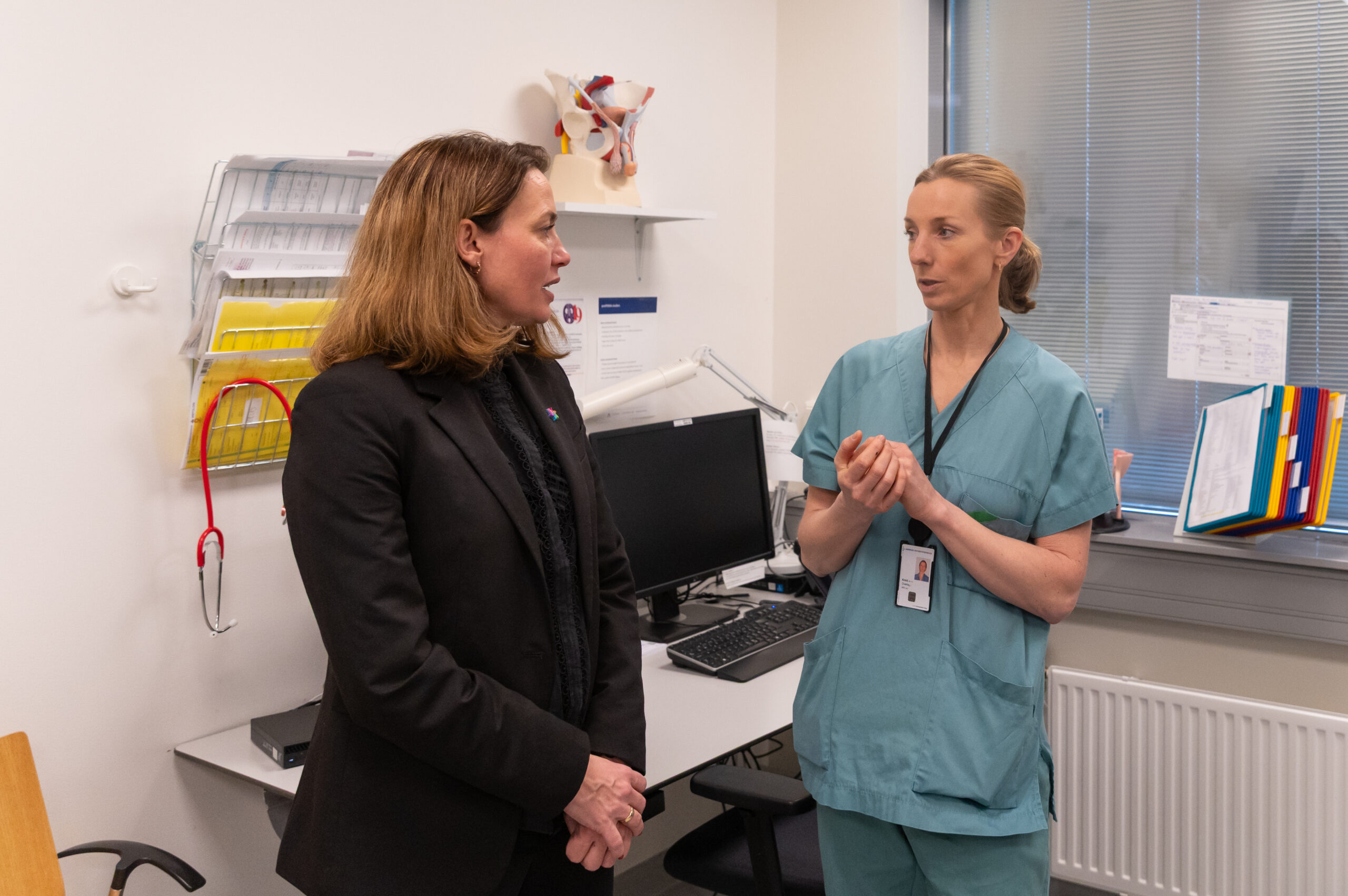To kvinner, den ene i svart dress og den andre i legekittel, fører en samtale på et legekontor med medisinsk utstyr i bakgrunnen.