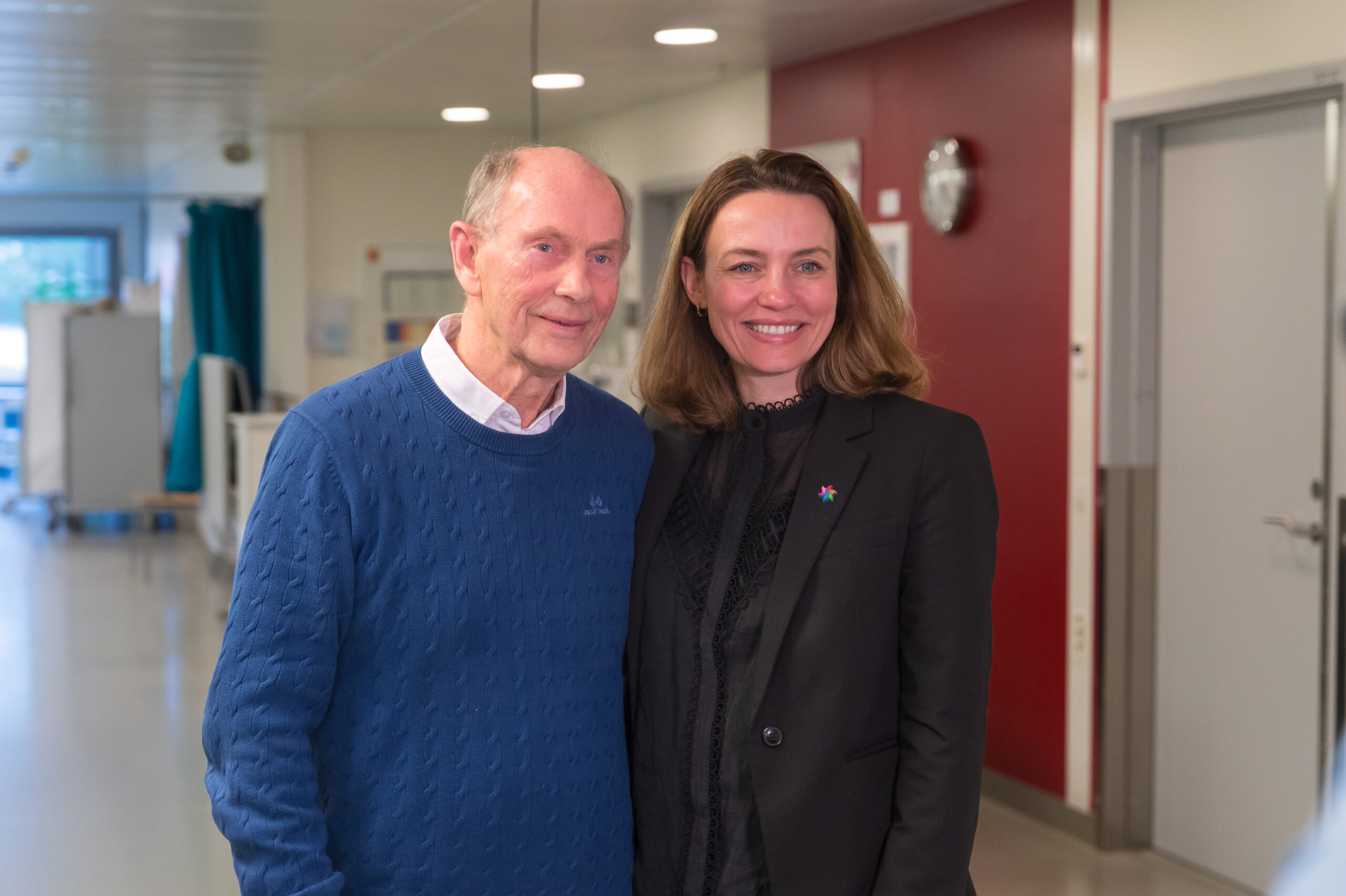 En eldre mann i blå genser og en kvinne i svart dress står sammen og smiler i en sterkt opplyst gang med røde og hvite vegger.