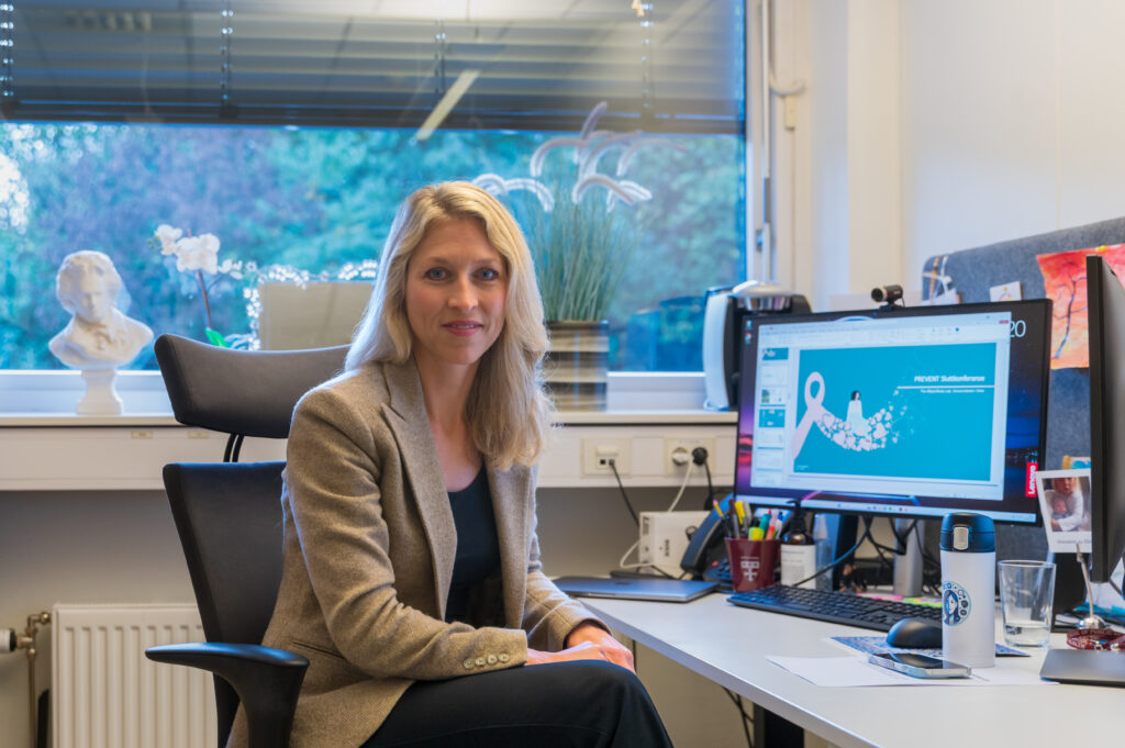 En kvinne med blondt hår sitter ved et skrivebord på et kontor, vendt mot kameraet. En dataskjerm viser en blå skjerm med et hvitt bevissthetsbånd.