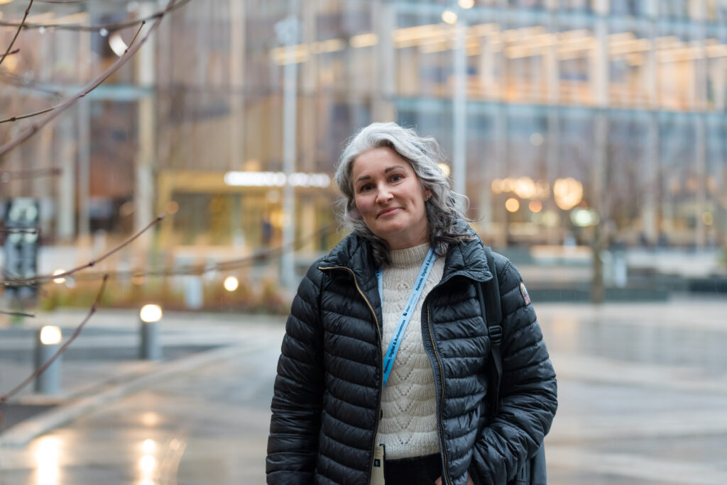 En kvinne med grått hår, iført svart jakke og nøkkelbånd, står utendørs i et urbant område med glassbygninger i bakgrunnen.