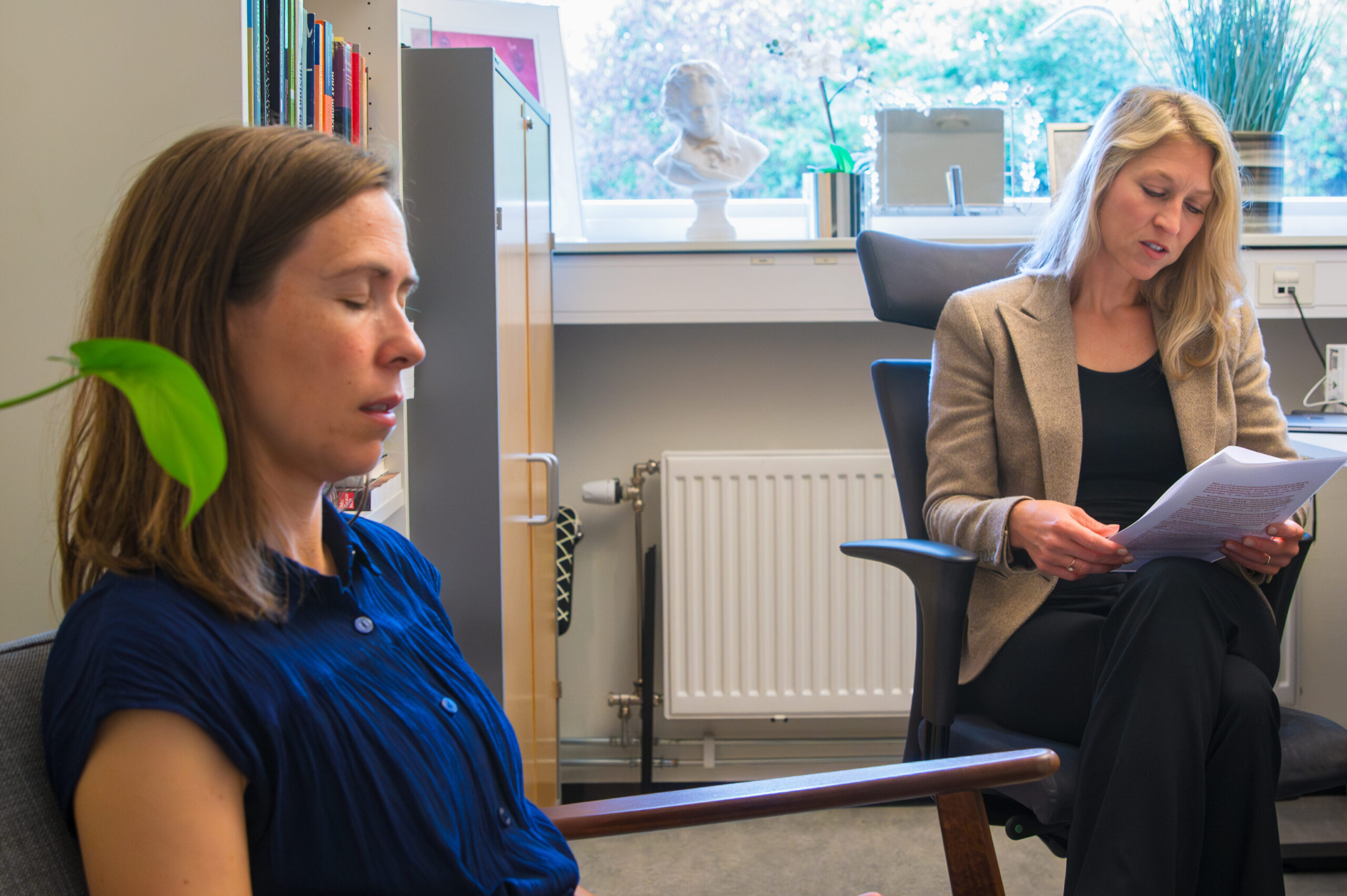 To kvinner sitter på et kontor; den ene sitter med lukkede øyne og hendene i fanget, mens den andre går gjennom dokumenter ved et skrivebord.